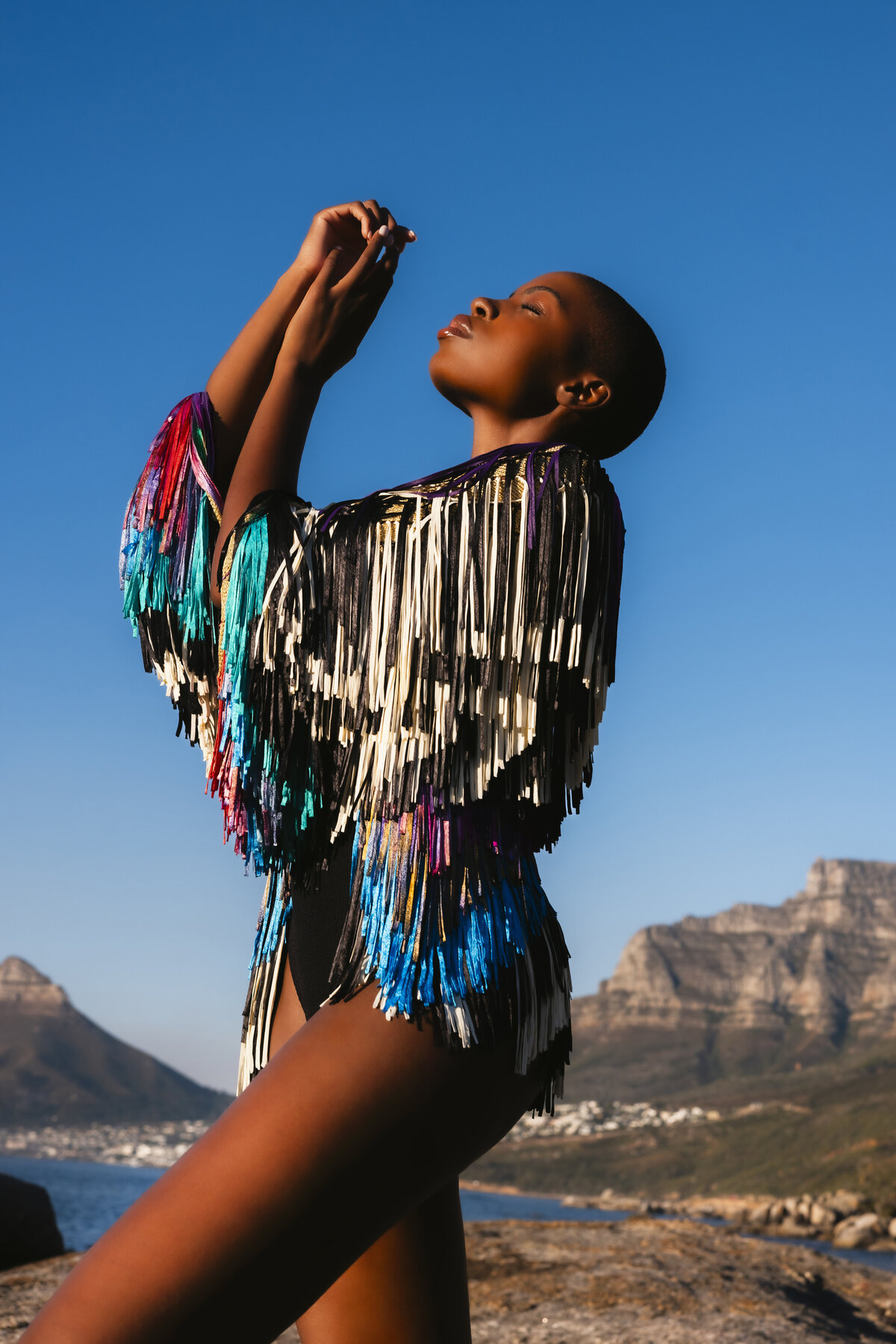 Model enjoying golden hour on the rocks in Cape Town, wearing a colorful fringe jacket over a black bodysuit, with clear blue skies and mountain views in the background.