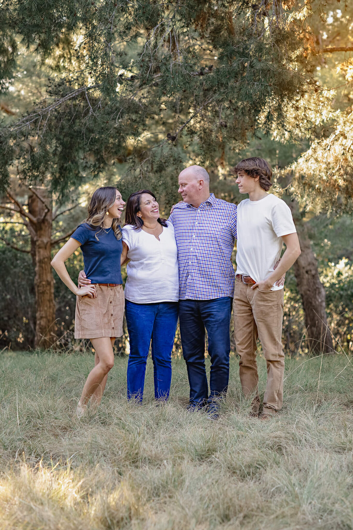 A family of four with dark hair standing in the grass in front of beautiful green trees, laughing and smiling at each other. Captured at Jeffrey Open Space in Irvine.