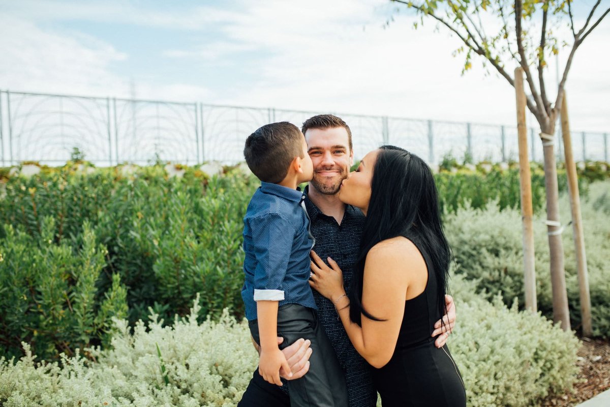 Father receives kisses on his cheek from his wife and young son amongst green shrubbery