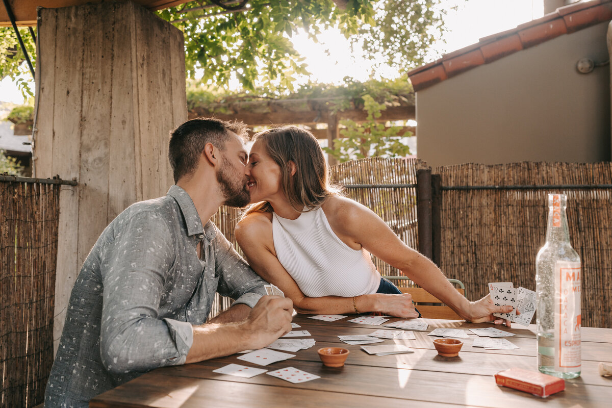 A photograph in color of Maggie and Caleb during their engagement session at Hotel San Jose in Austin, Texas. The couple are seated outdoors at a table with a card game of rummy in progress and a bottle of mezcal opened with clay mezcal cups. They are leaning towards each other and kissing. Wedding photography by Stacie McChesney/Vitae Weddings.