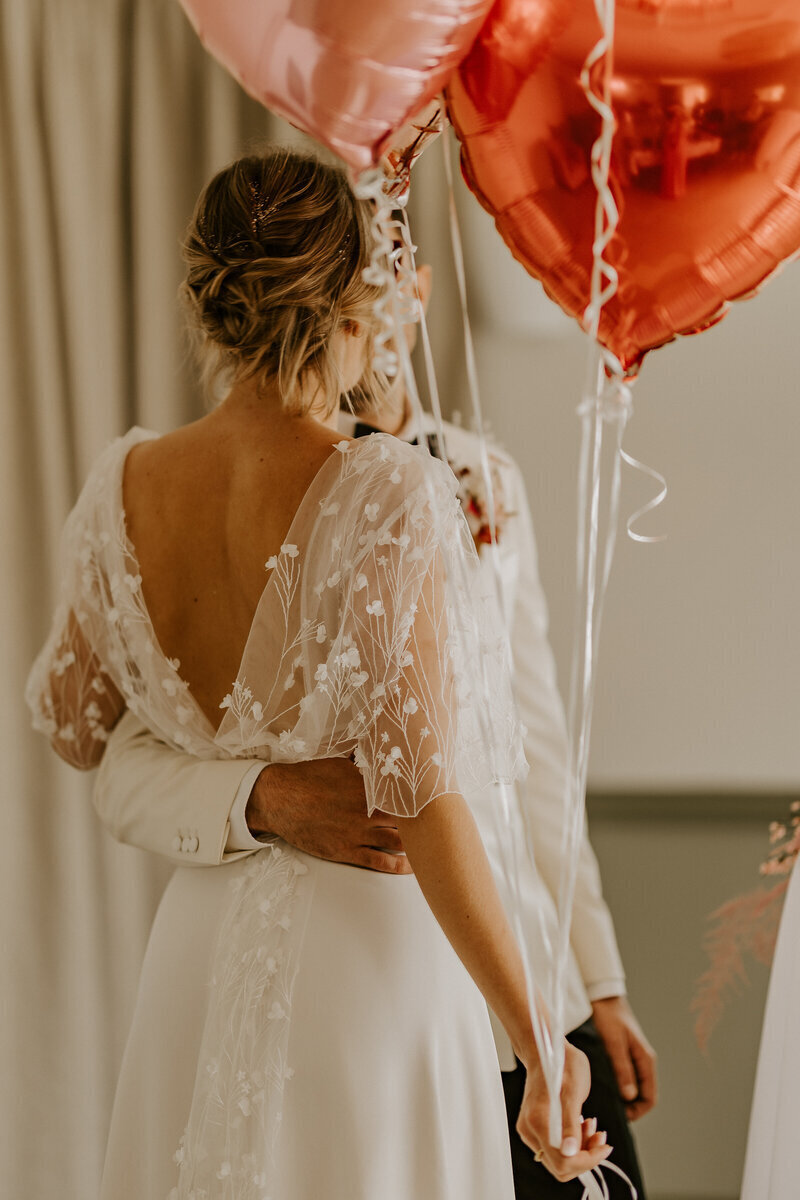 Mariée de dos tenant dans ses bras son mari dont on n'aperçoit pas le visage. Décor de ballons métalliques coeur rouge et rose. Séance photo pour la workshop photographie de mariage.