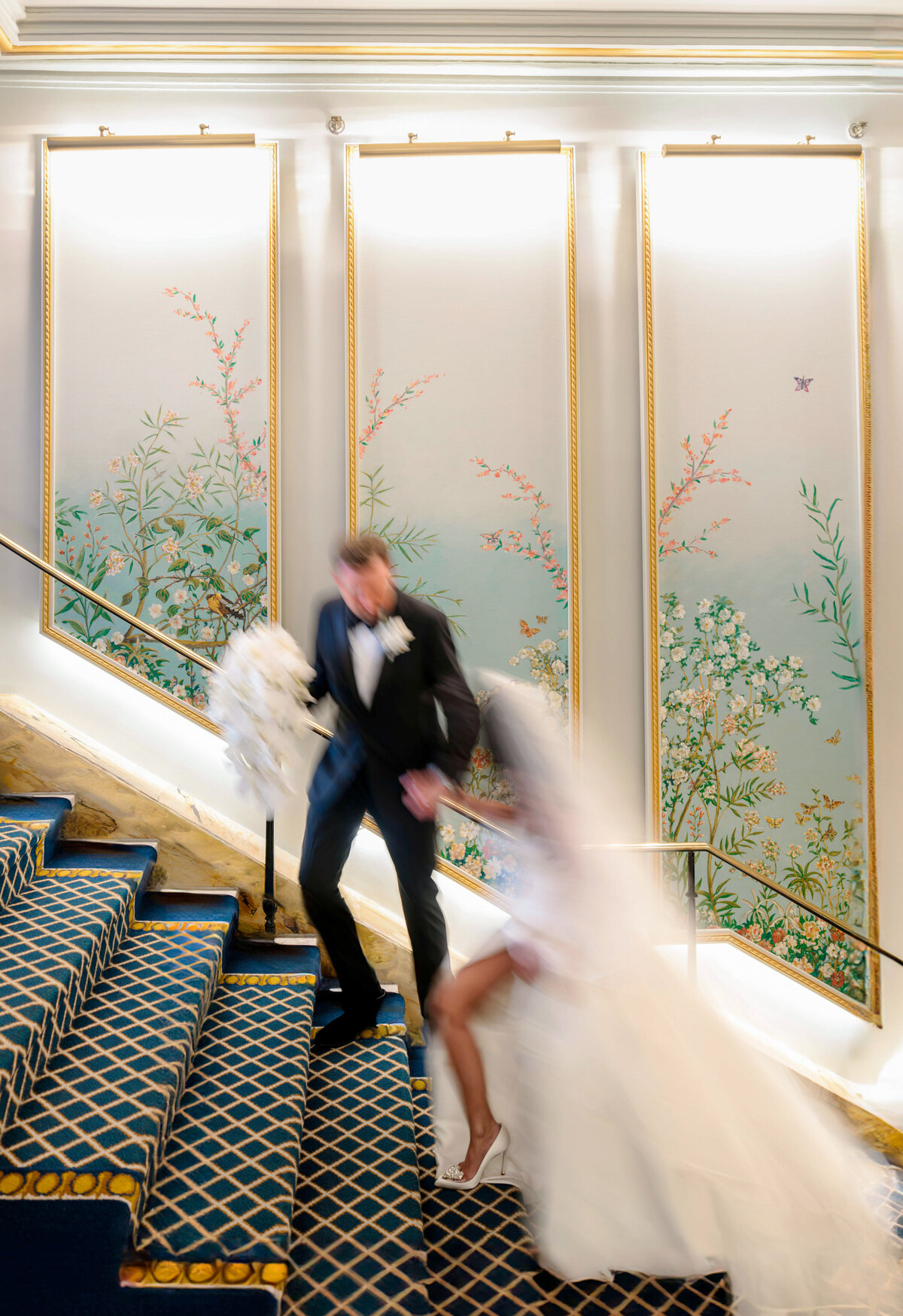 A bride and groom descend a staircase adorned with a blue and gold carpet. The walls are decorated with elegant floral panels. The brides long veil flows behind, and the groom holds her hand while carrying a bouquet of white flowers.