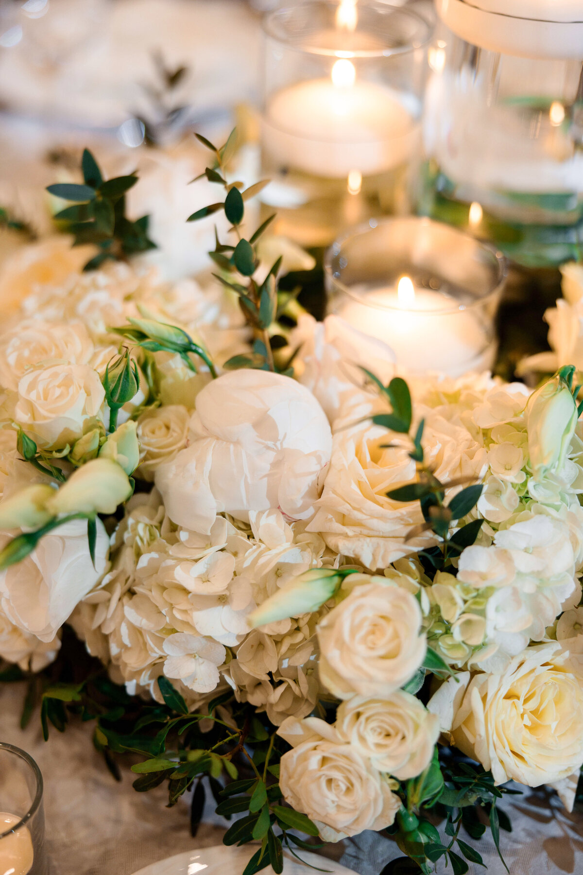 A lush floral centerpiece featuring white roses, peonies, and hydrangeas, accented with greenery. Surrounding the flowers are clear glass votives with lit candles, setting a warm and elegant atmosphere on a table.