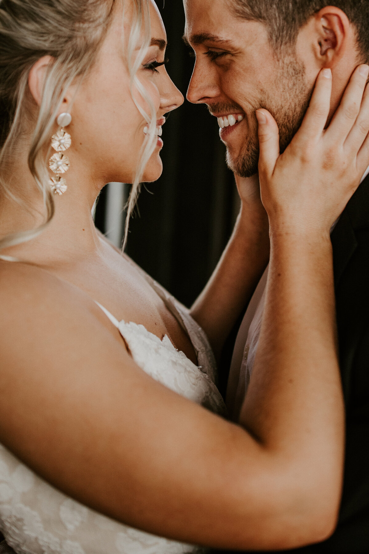 A smiling bride wearing a white wedding gown and earrings holds a smiling groom by the jawline while locking eyes.