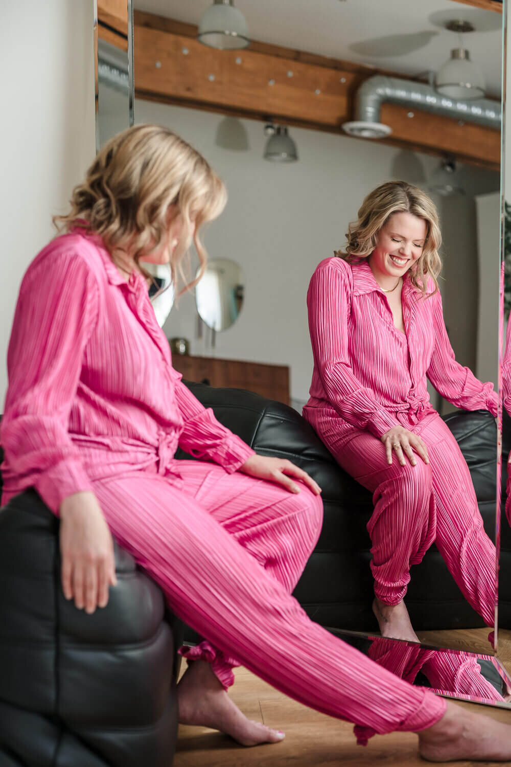 Naturopathic physician in pink pleated outfit smiling while seated, reflected in tall mirror.