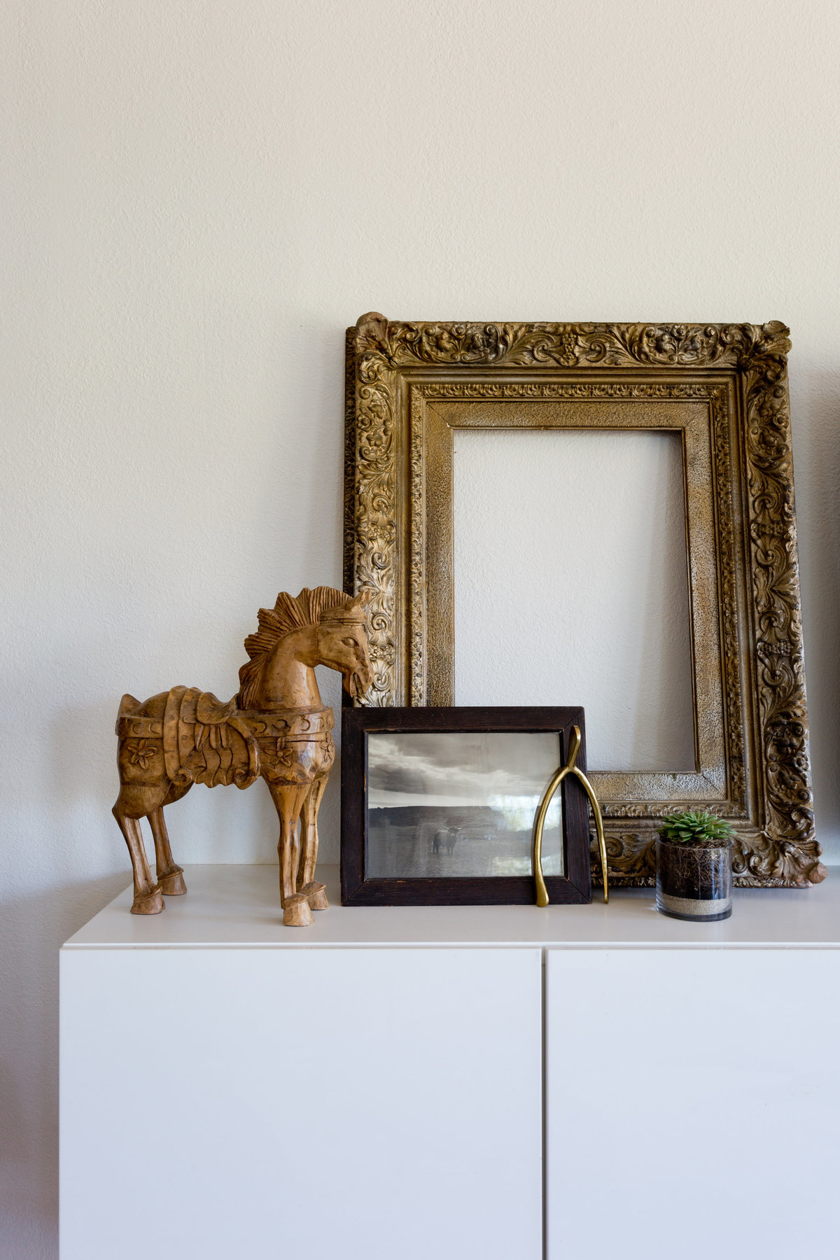 Styling of media cabinet with wooden horse, vintage gold frame, black and white photo of short horn cow, gold wishbone, and succulent