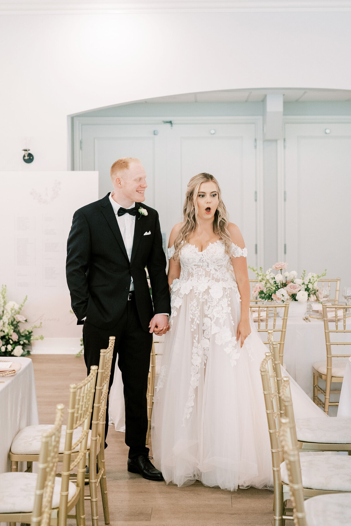 A bride and groom stand together indoors, dressed in wedding attire. The bride wears a white gown with lace details and an off-the-shoulder design, while the groom wears a black tuxedo. They both look surprised at their modern &amp; romantic wedding in Calgary at Norland Historic Estate.