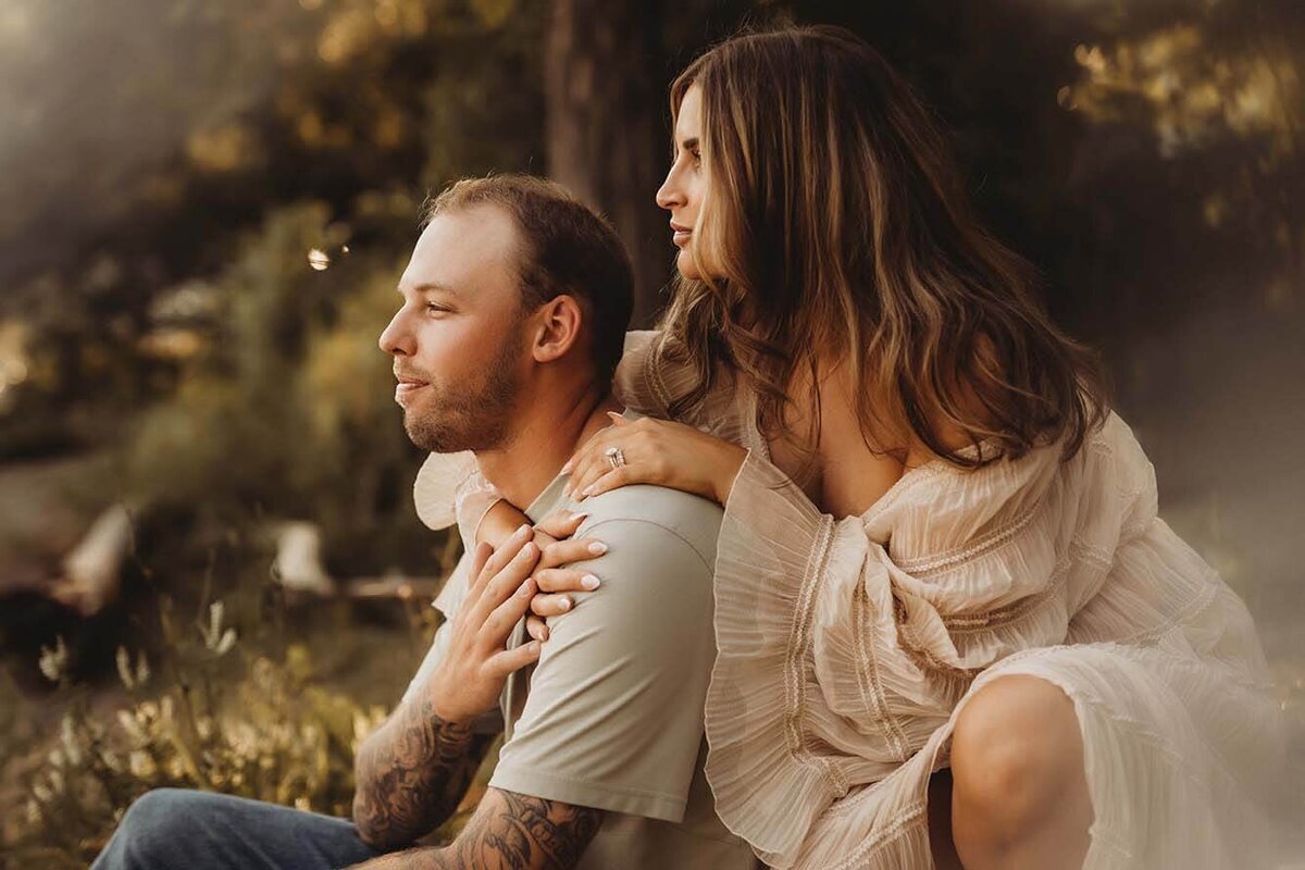a dad sits on the grass while his wife crouches behind him with her arms wrapped around him and his hands on her hands