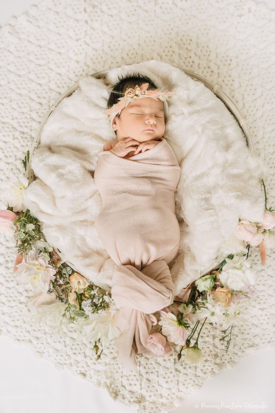 Full floral basket – Newborn baby girl swaddled in blush pink, sleeping in a round basket lined with fluffy white fur and surrounded by pastel flowers.