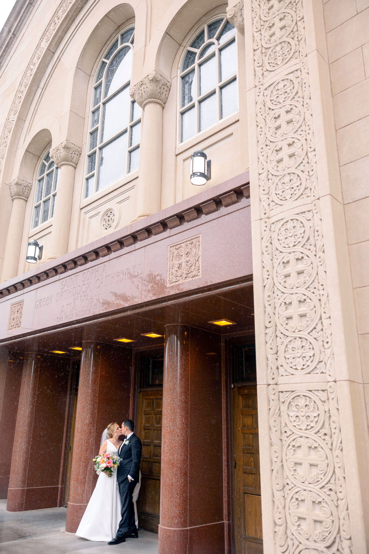 A bride and groom stand in front of an ornate building entrance, holding a bouquet. The building features tall arched windows, intricate decorative stonework, and a sign reading Greek Orthodox. The couple shares a moment under the overhang.