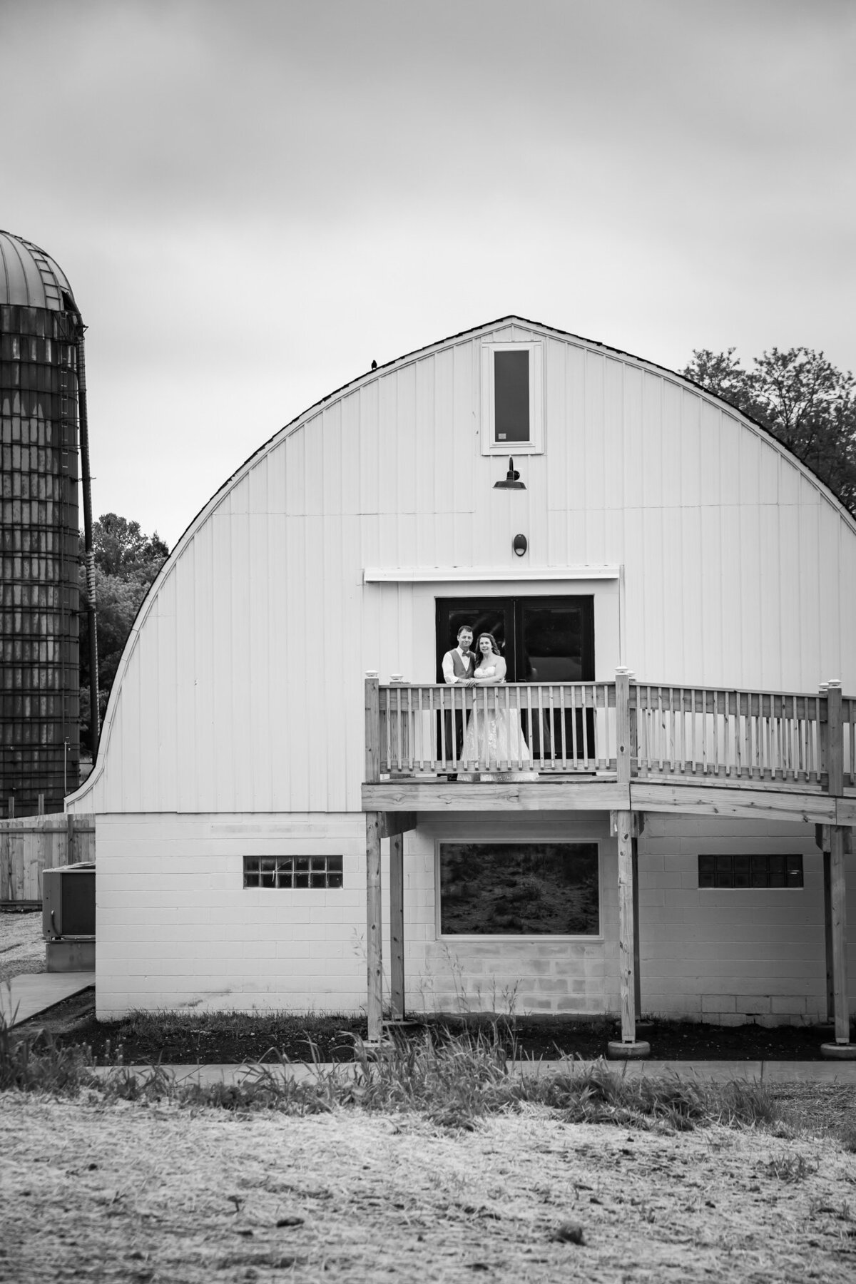 bride and groom stand on deck in front of barn in black and white