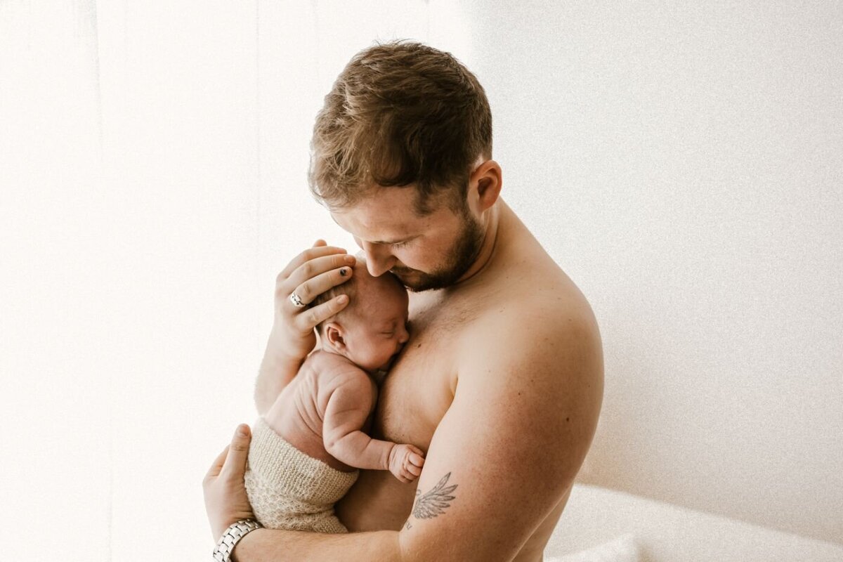 A touching image of a father cradling his newborn baby, who is wrapped in a soft knit, against his bare chest. Newborn photography by Ruaa Hamdan