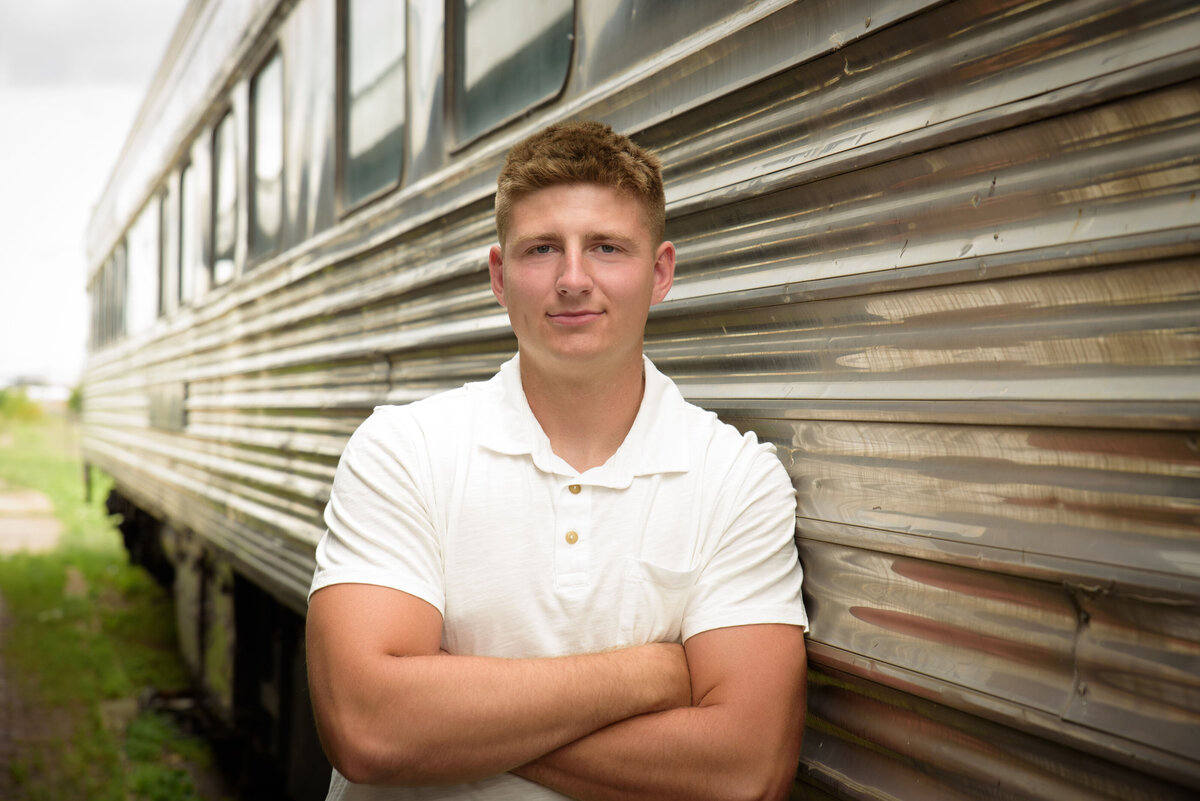 De Pere High School senior boy wearing blue shorts and a cream hooded sweatshirt in front of a train car at The Depot in Downtown Green Bay, Wisconsin