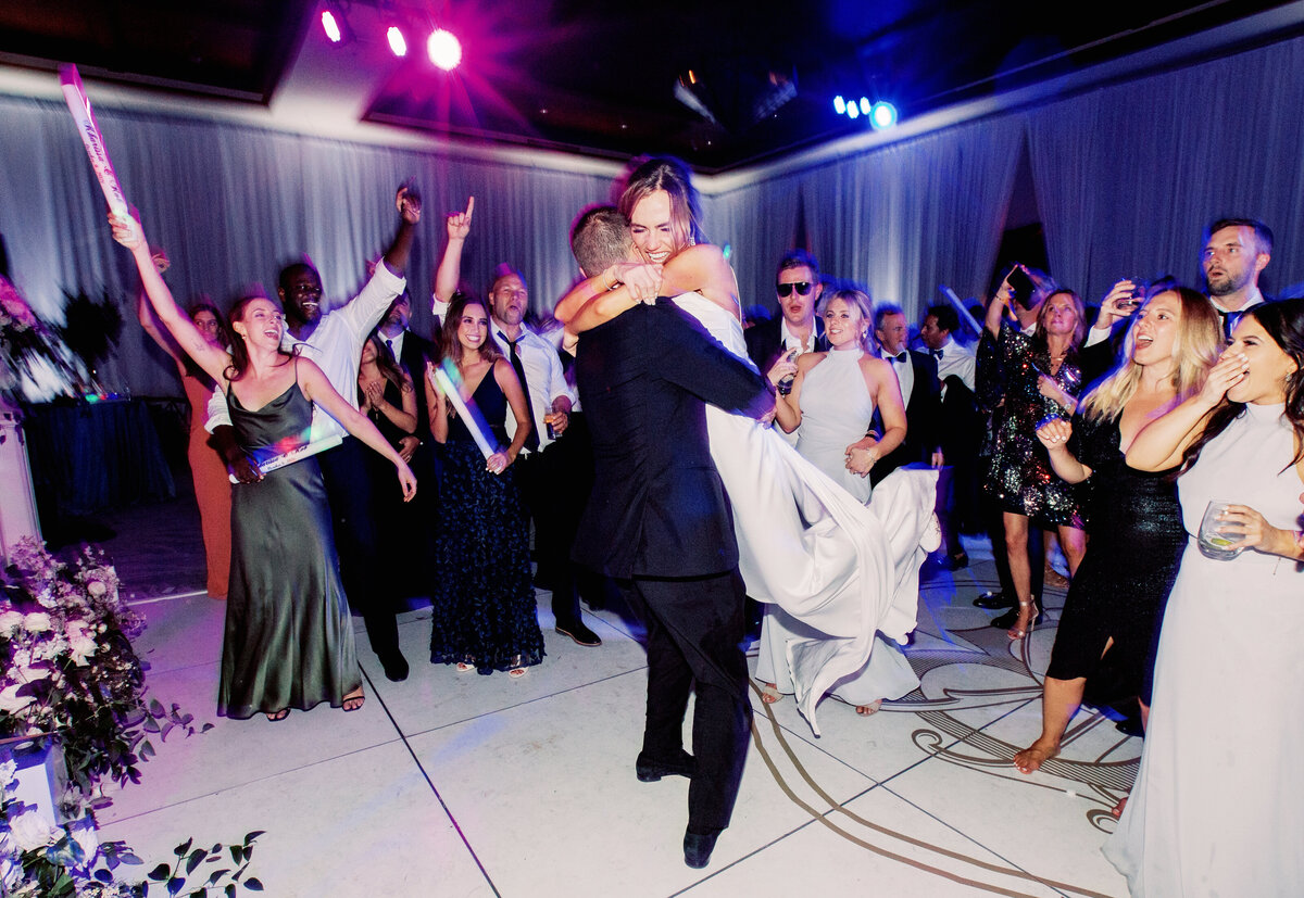 The bride and groom share an energetic dance floor moment, surrounded by guests celebrating with glow sticks and cheers.
