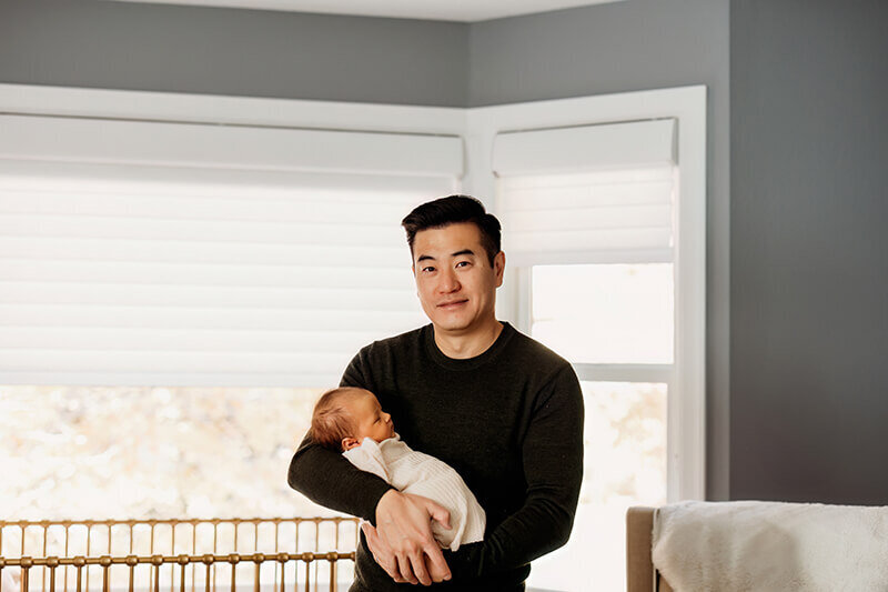 Dad smiles at camera while holding newborn baby girl in nurery