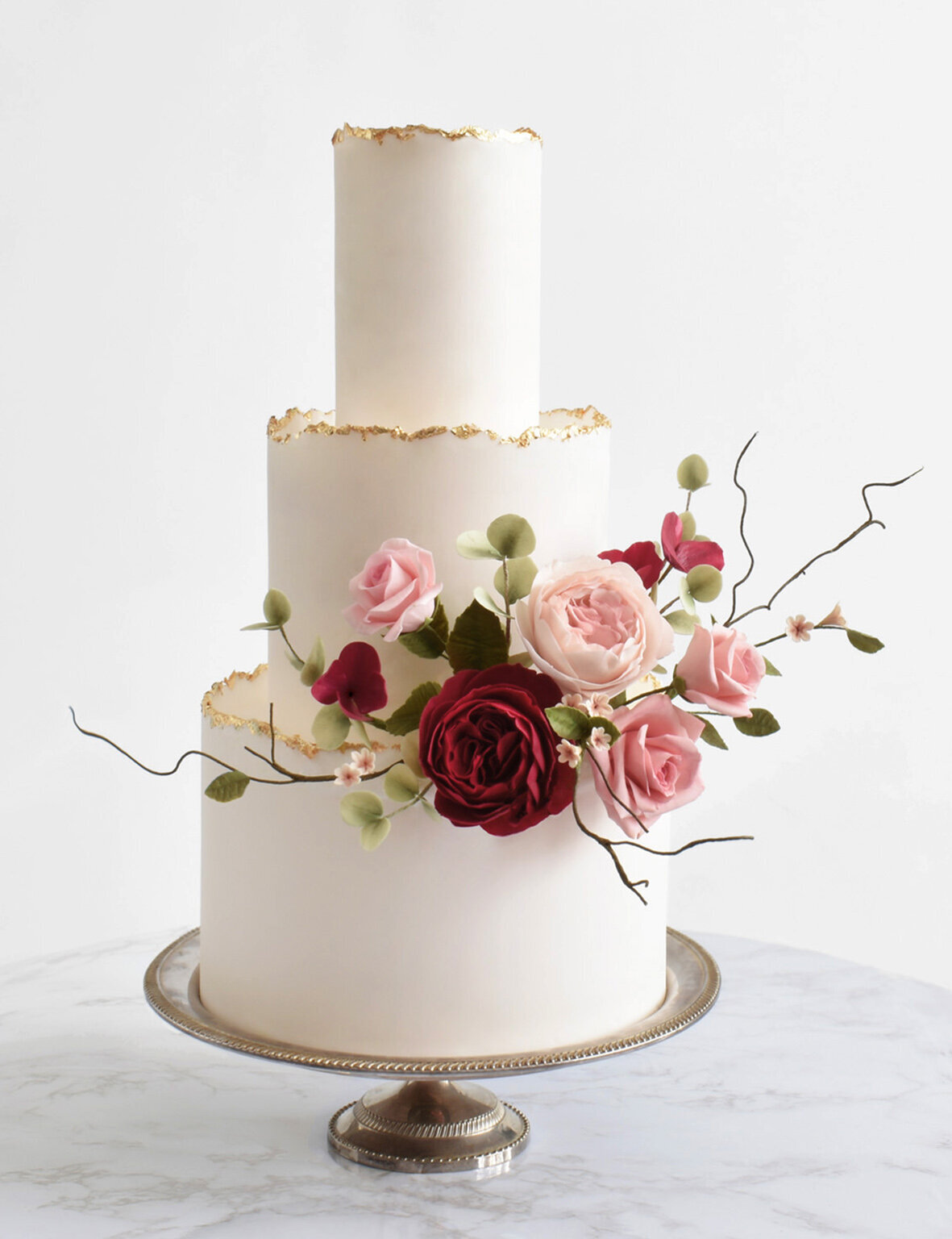 a three tiered white wedding cake with gold leaf edged and a bouquet of pink and deep red roses made out of sugar
