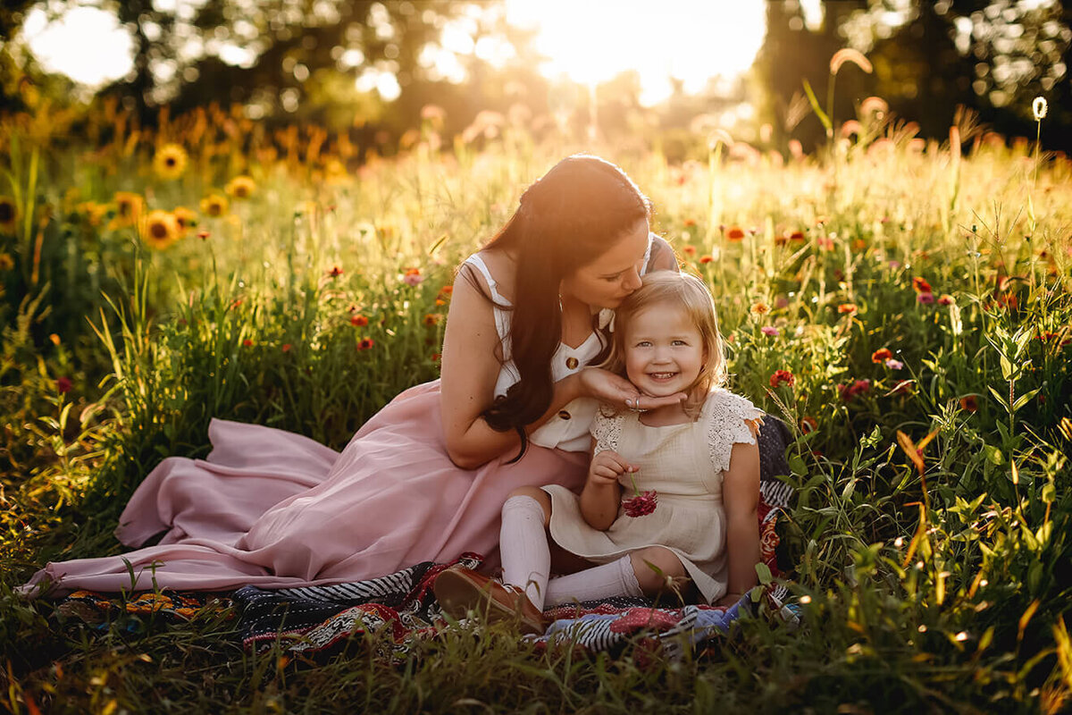 Mother and daughter sitting in flowers kissing little girls head