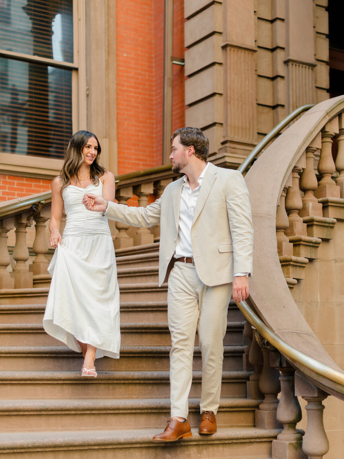 A couple descends the grand staircase outside The Union League of Philadelphia, exuding timeless elegance and connection. The groom-to-be reaches for his partner’s hand as she gracefully lifts her white dress, framed by the historic red brick and stone architecture. Captured by Elizabeth Kane Photography, a Luxury Philadelphia Wedding Photographer.