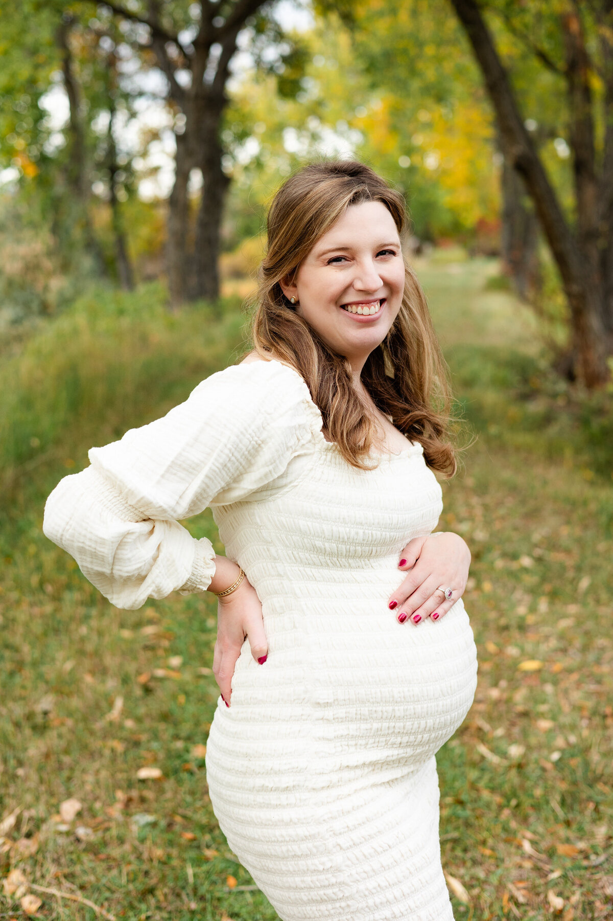 A pregnant woman has one hand on her back and one hand on her belly as she smiles at the camera.