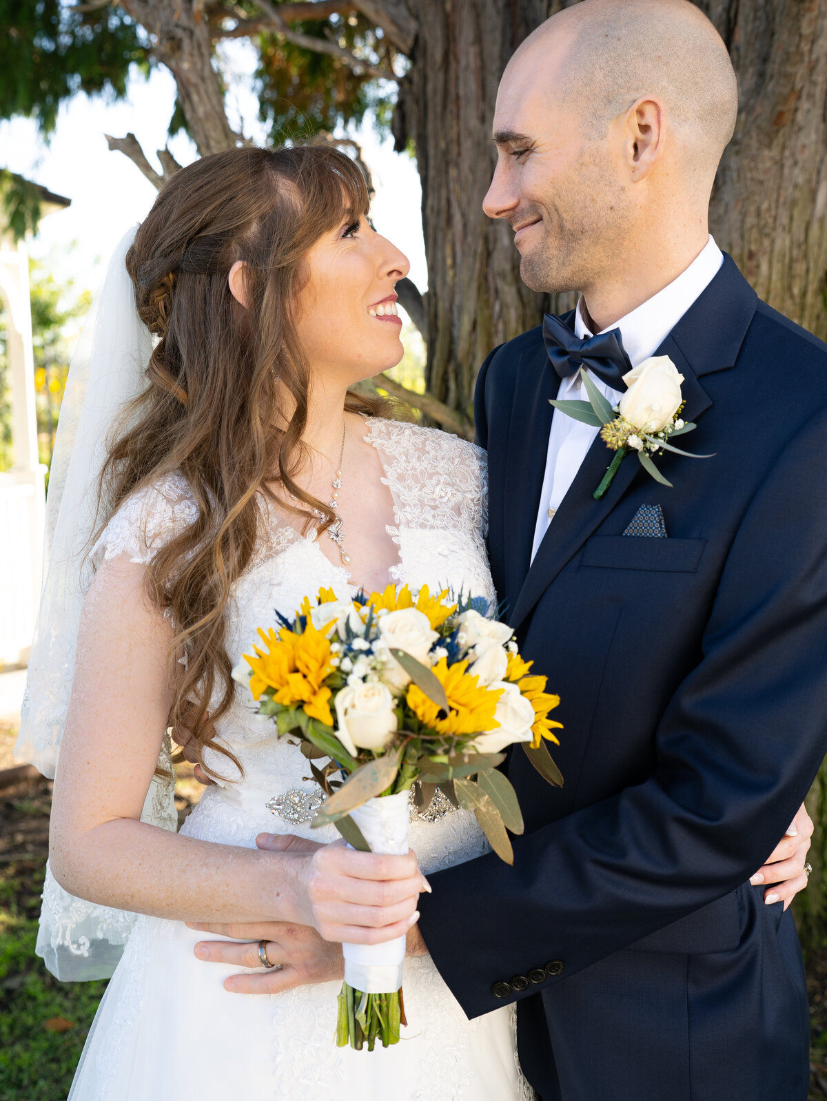 A bride with long red hair and lace cap sleeves holds a sunflower bouquet in one hand wraps her other arm around her groom as he pulls her close and they face each other with a sweet smile. Photo by SAVI Photography - San Diego California Photographer