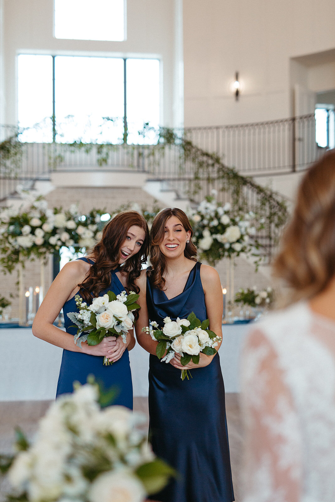 Two bridesmaids in blue satin gowns holding white bouquets smile in front of a table with tall white florals.