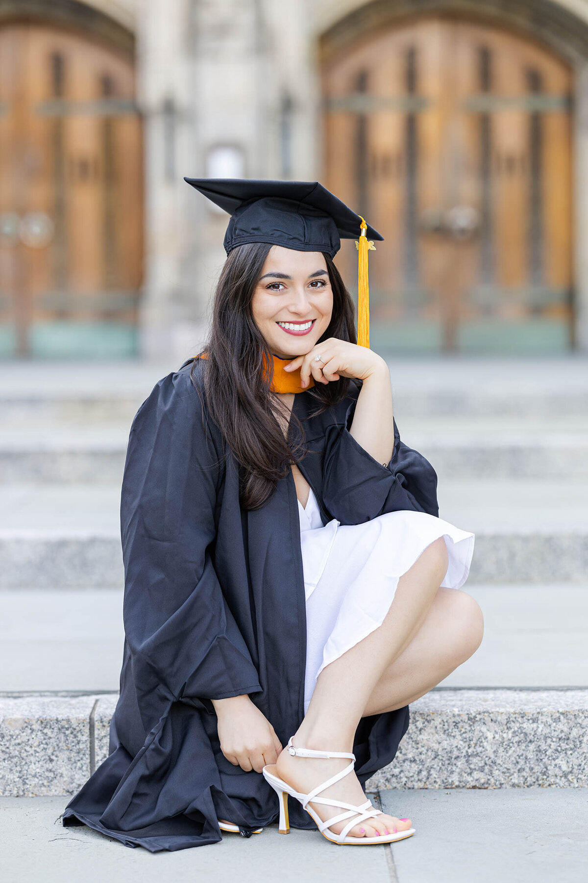 Graduate girl wearing cap and gown sits on the steps as she stairs smiling at the camera during her graduate photo session in Connecticut