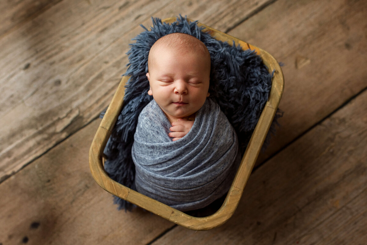 newborn-boy-swaddled-in-blue-on-wood-floor-backdrop-in-york-pa-newborn-studio
