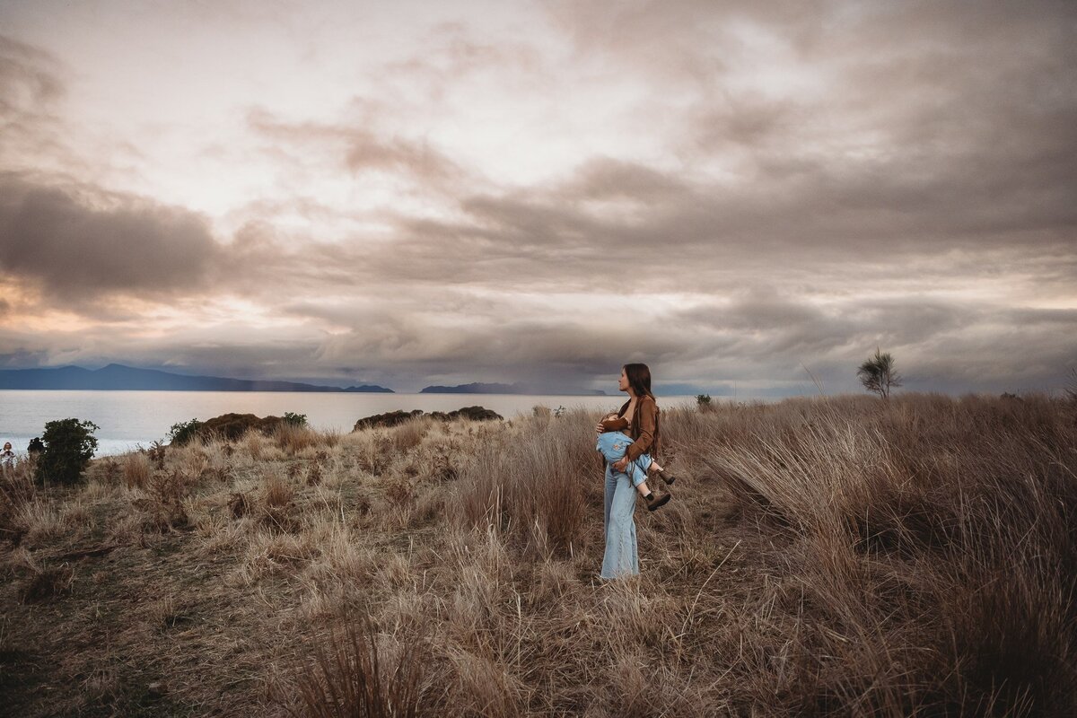 A person stands in a grassy field during sunset, holding a newborn. They both face towards a distant view of the sea and cloudy sky, capturing the essence of Wollongong motherhood sessions.