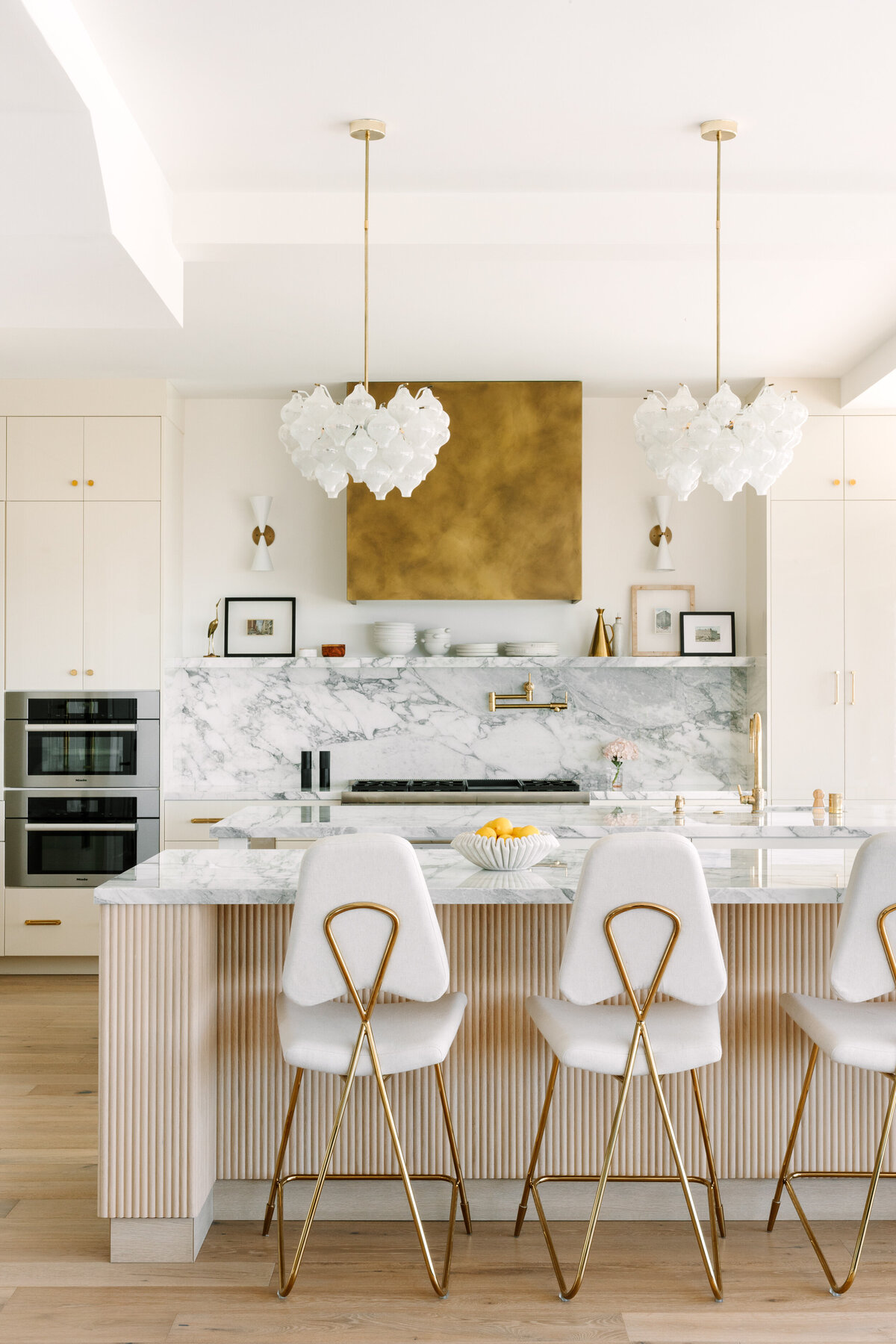 Kitchen with high gloss cream cabinets and Vagli marble counter and backsplash, brass range hood, Thermador appliances and vintage 1960's pendants.