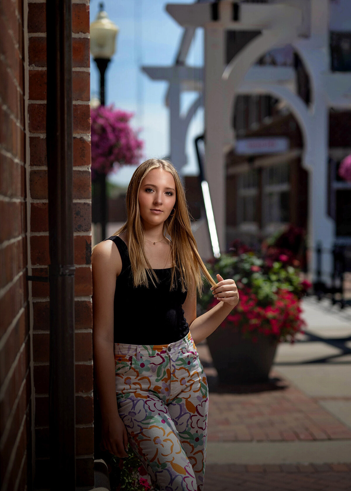 A high school grad leans on a brick wall twirling her hair in colorful pants