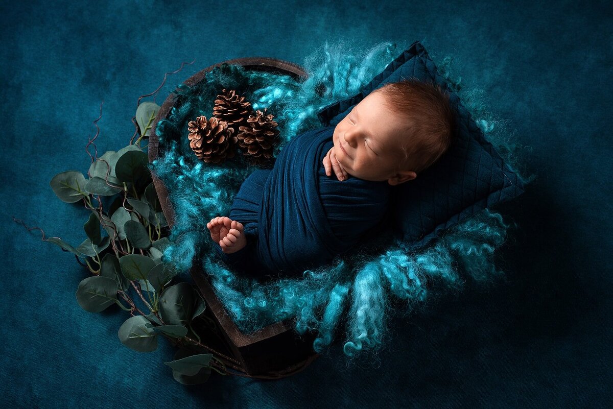 Smiling newborn boy wrapped in teal posed in a heart basket