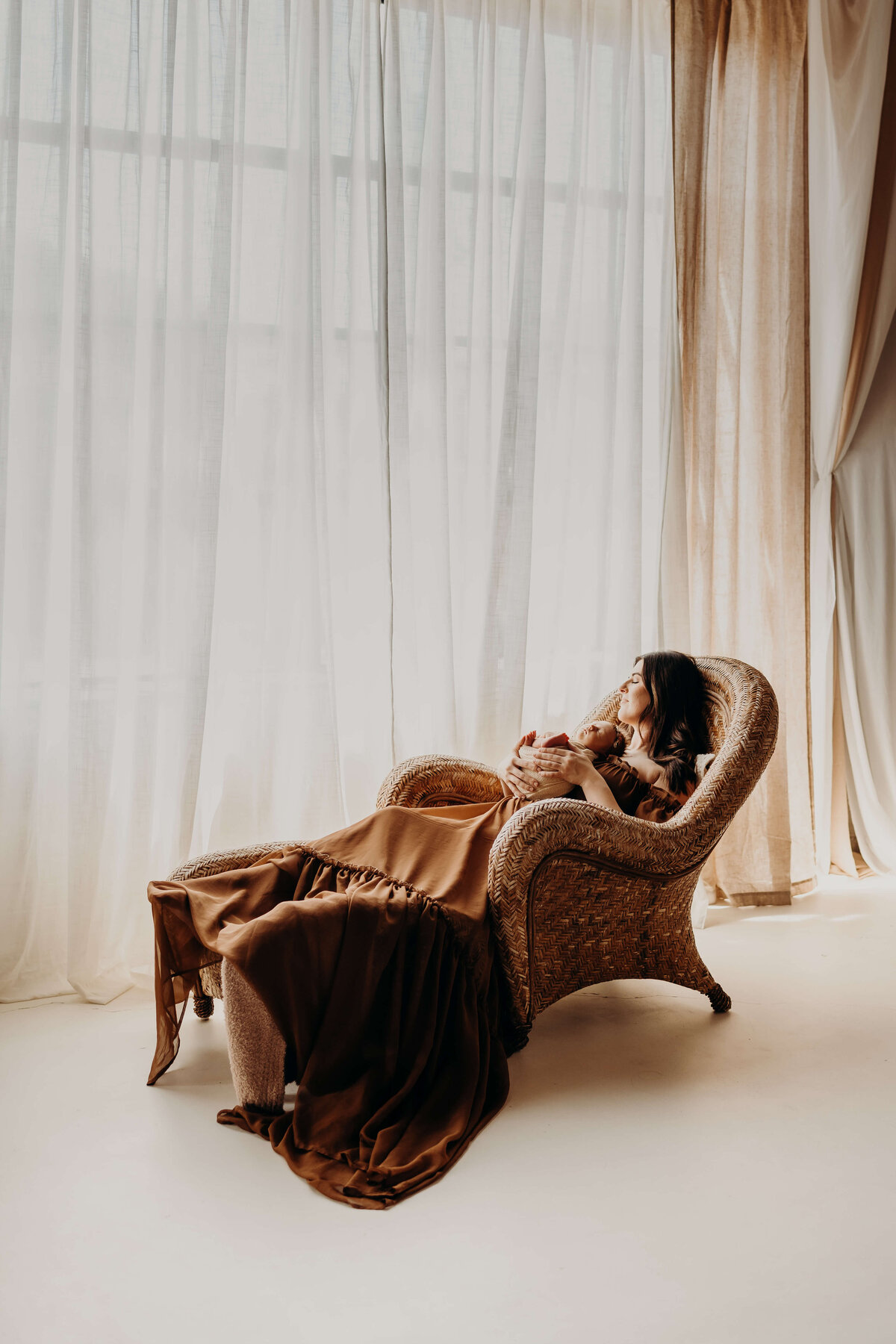 newborn mother sits in a high back wicker chair reclining next to large windows