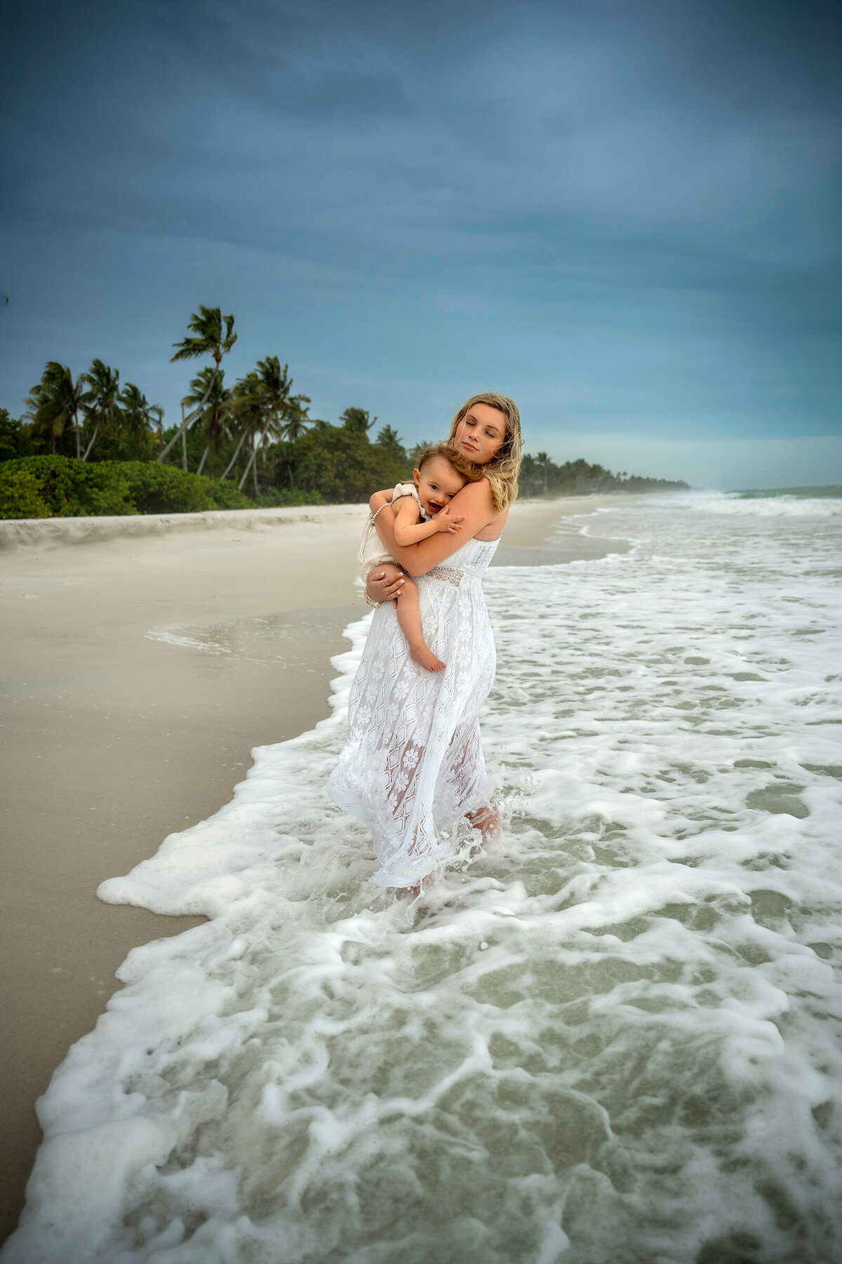 Pregnant mom holding daughter dressed in white during the storm  in the waves at Naples beach
