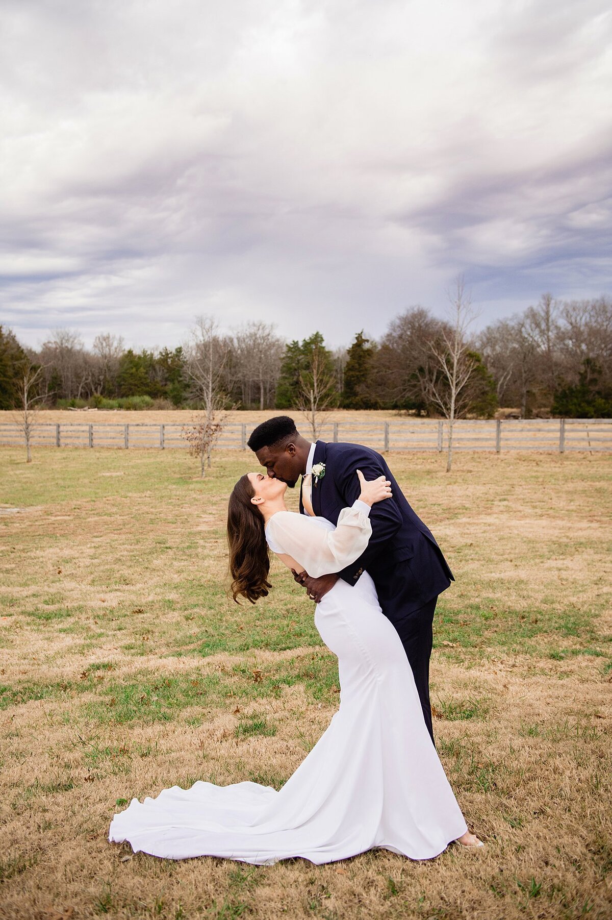 The bride, wearing a fitted wedding dress with a flowing train and sheer long sleeves bends back and the groom dips her into a deep kiss. The groom is wearing a black suit with a white shirt and white boutonniere. They are standing in a field at Steel Magnolia Barn.