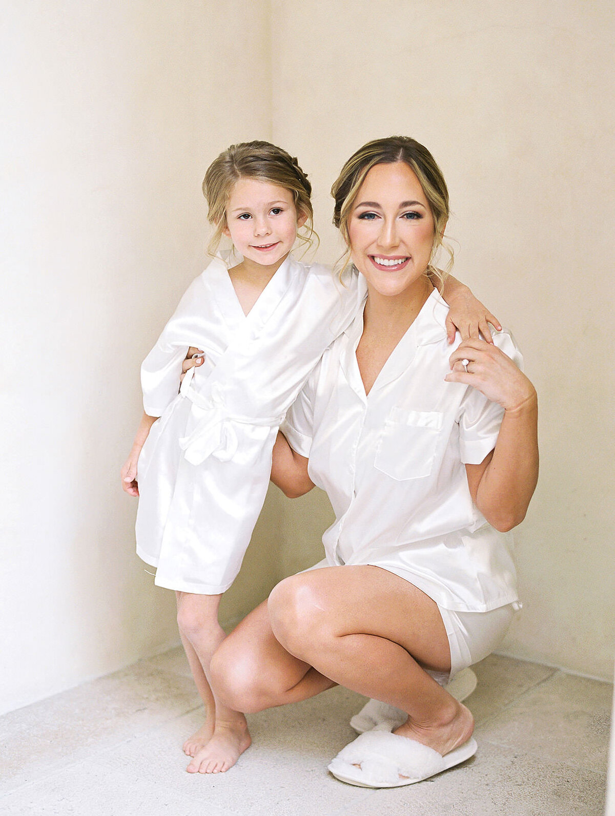 A bride and her flowergirl pose together in pajamas against a neutral wall