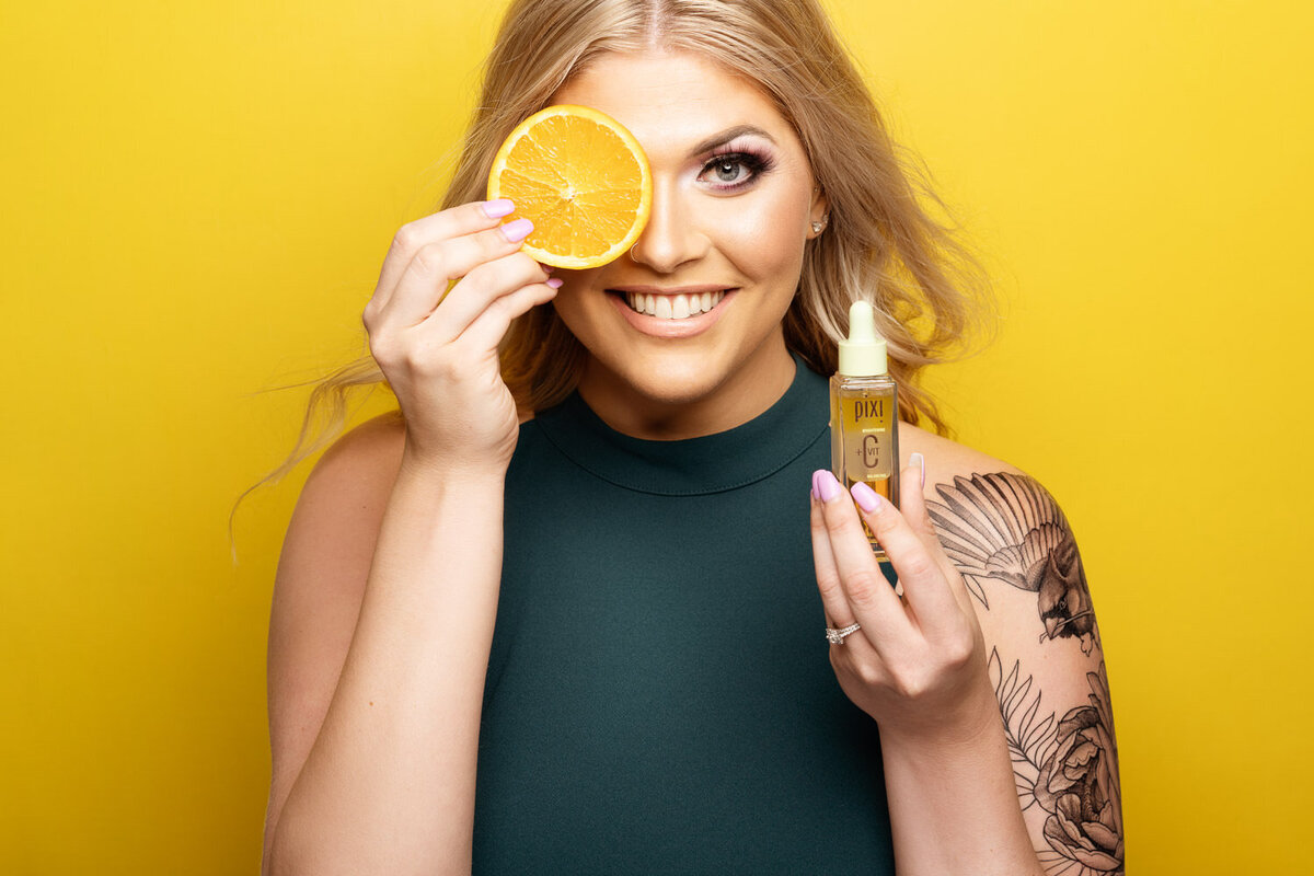 Studio branding photo of a woman in a green dress holding a skincare product and an orange slice against a yellow backdrop, representing natural ingredients. Photographed in Austin, TX.