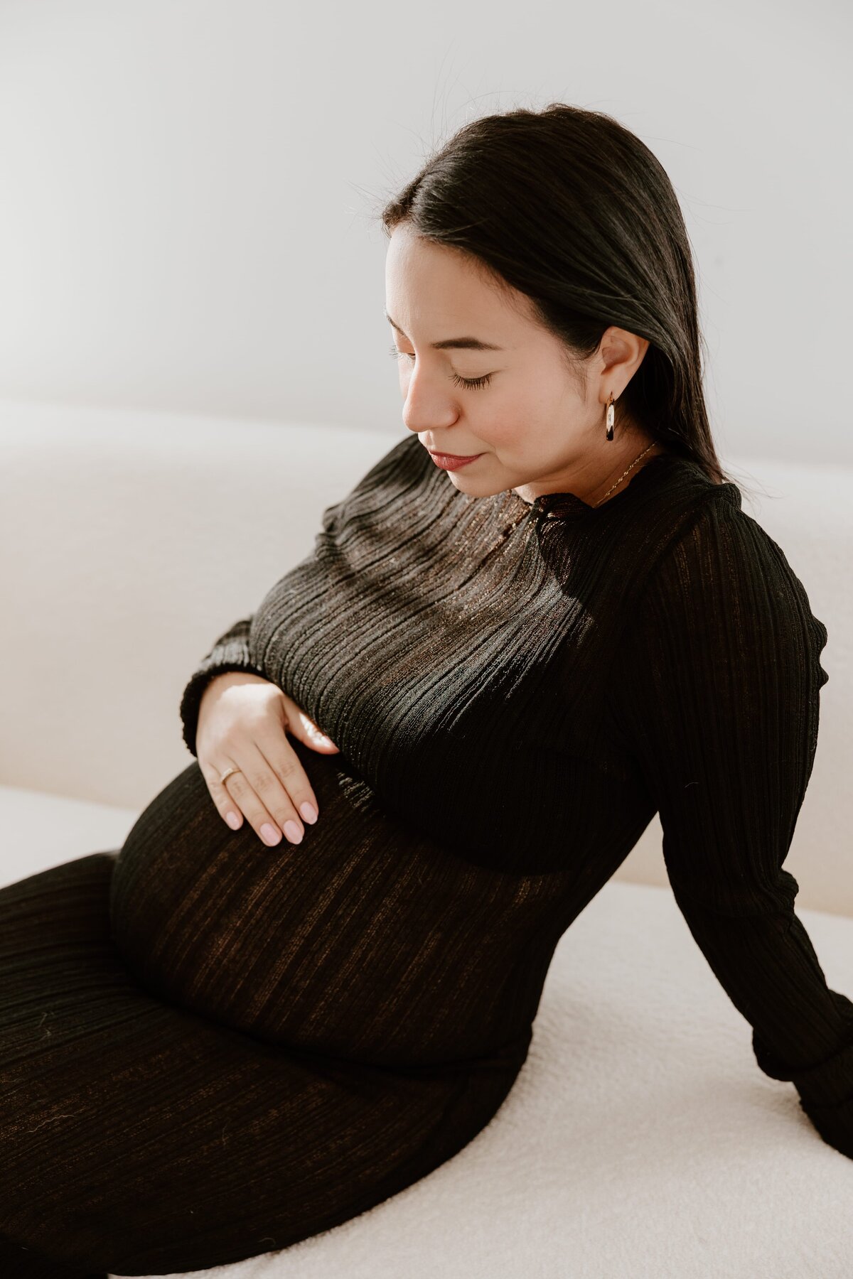 A pregnant woman with dark hair sits on a light-colored couch, wearing a black, ribbed, long-sleeve dress. She gently cradles her baby bump with one hand while looking down with a soft, peaceful expression. Sunlight highlights her face and the texture of her dress, creating a warm, intimate atmosphere.