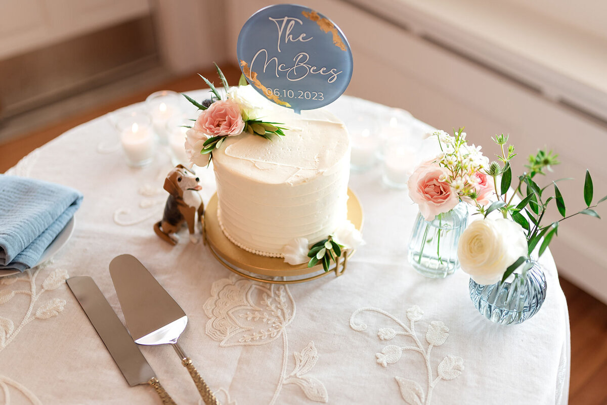 A wedding cake table with a simple white cake topped with flowers, a cake knife set, and a sign with the couple's surname and wedding date.