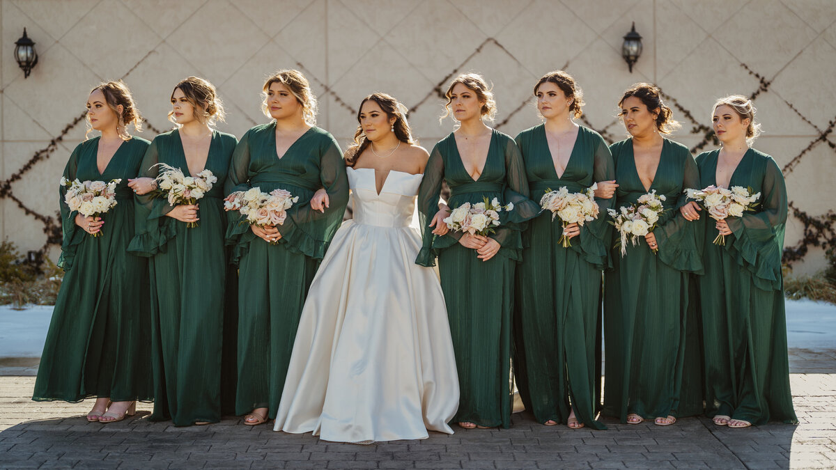 Elegant group photo of a bride and her bridesmaids in stylish emerald green dresses, locking arms and looking in the distance as they take in the moment against an Italian-inspired tiled plant wall at the D'Vine Grace Vineyards wedding venue in DFW, Texas