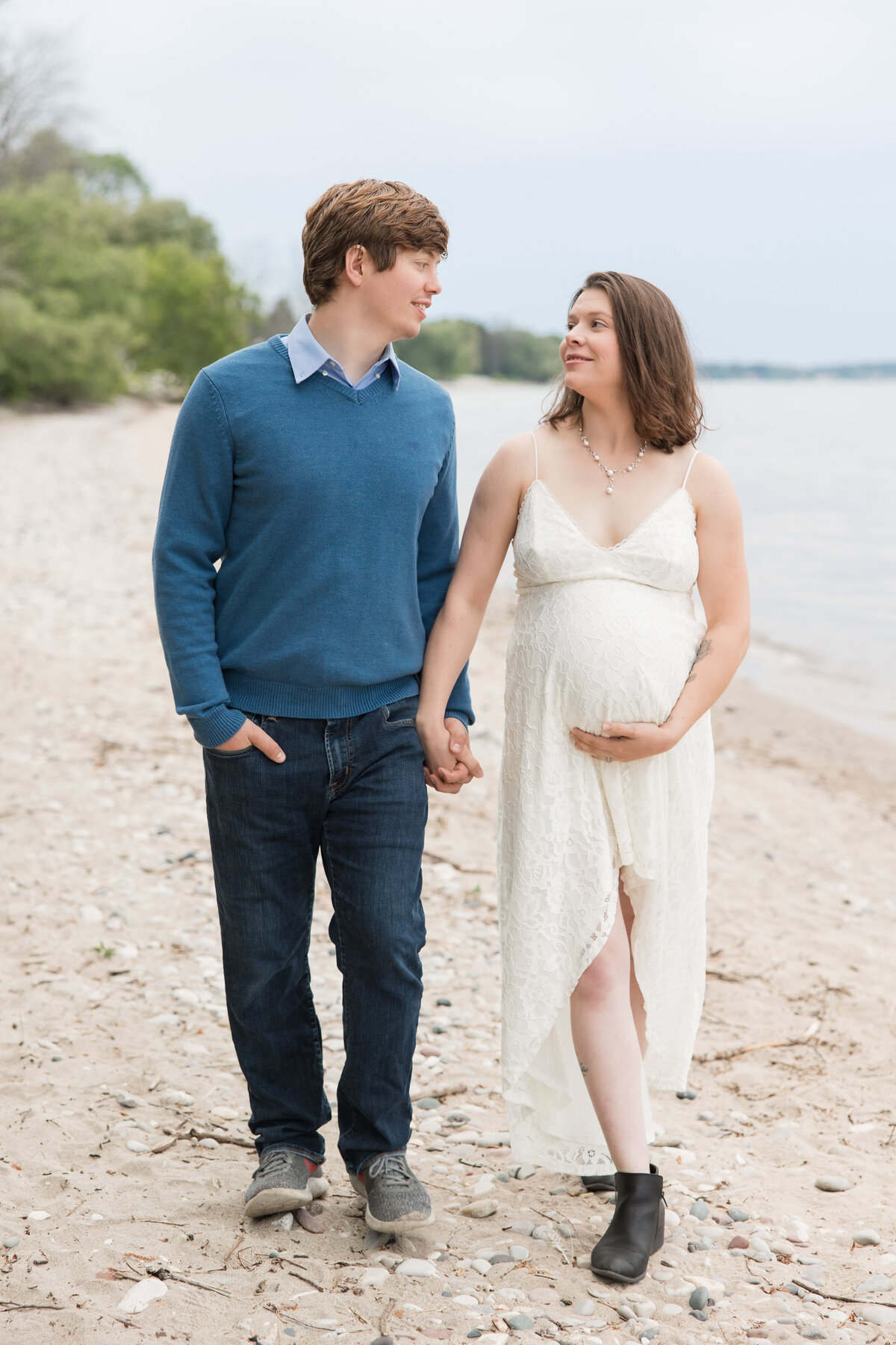 Expectant couple walking along the beach, holding hands, and smiling at each other