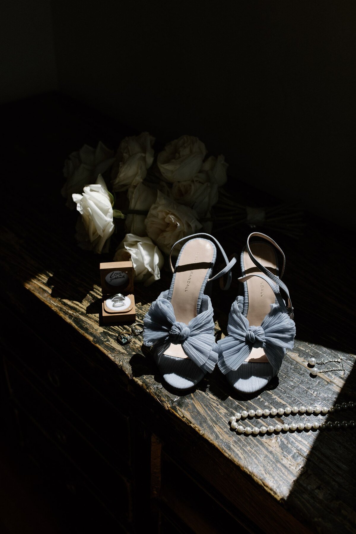 Flat lay of bridal details: blue bow heels, pearl necklace, wedding rings in a wooden box, and white rose bouquet on a rustic wooden dresser.
