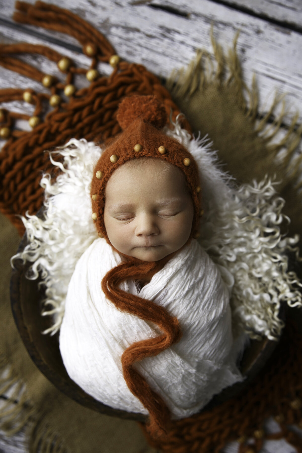Small newborn baby laying in a basket with an orange colored cap and orange knit scarf underneathe him