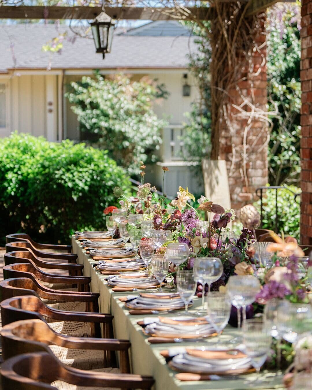 An elegant outdoor dining setup with a beautifully decorated long table adorned with fresh florals and fine glassware under a pergola. 