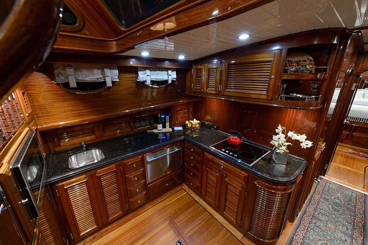 Elegant yacht kitchen featuring polished wooden cabinetry, black granite countertops, stainless steel appliances, a built-in stove with a red pot, a sink, flowers, and various kitchen items
