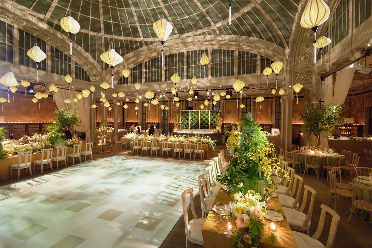 The elegant and spacious wedding reception hall, adorned with numerous hanging lanterns and lush floral arrangements, is located at the New York Public Library in New York City. The captivating setting was beautifully captured by photographer Shawn Connell. Videographers Aaron Navak Films also expertly documented the event, ensuring all the precious moments were preserved for years to come. Long dining tables are elegantly decorated with white linens, candles, and stunning flower centerpieces, creating a romantic ambiance for guests to enjoy. The central area features a large and well-lit dance floor, inviting guests to join in the celebration and create unforgettable memories.