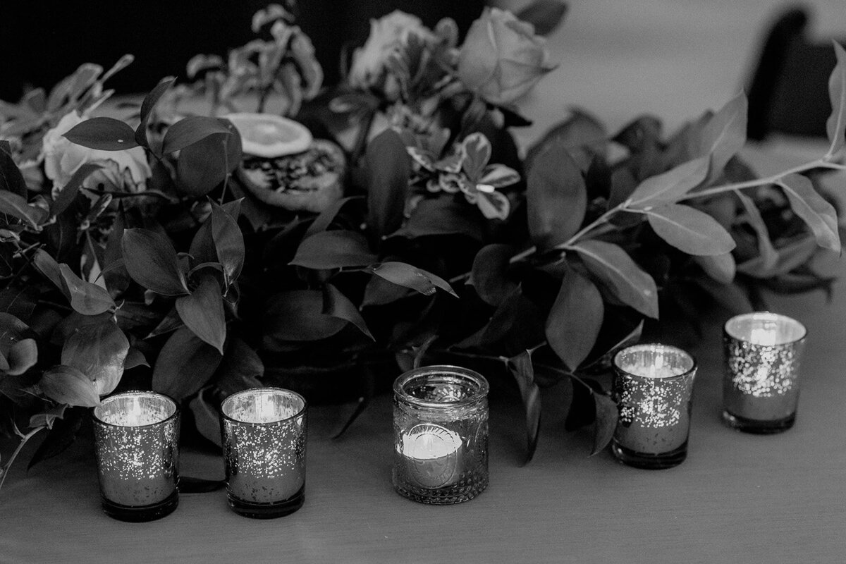 A black and white photo of a floral arrangement with greenery and roses, lined with glittering tealight candles in small glass holders