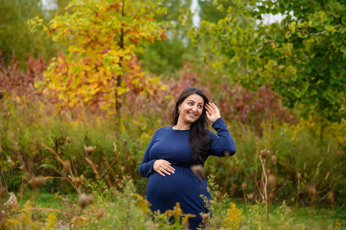 woman with her hair in her hand wearing a blue maternity dress for her fall outdoor maternity photography session in Burlington