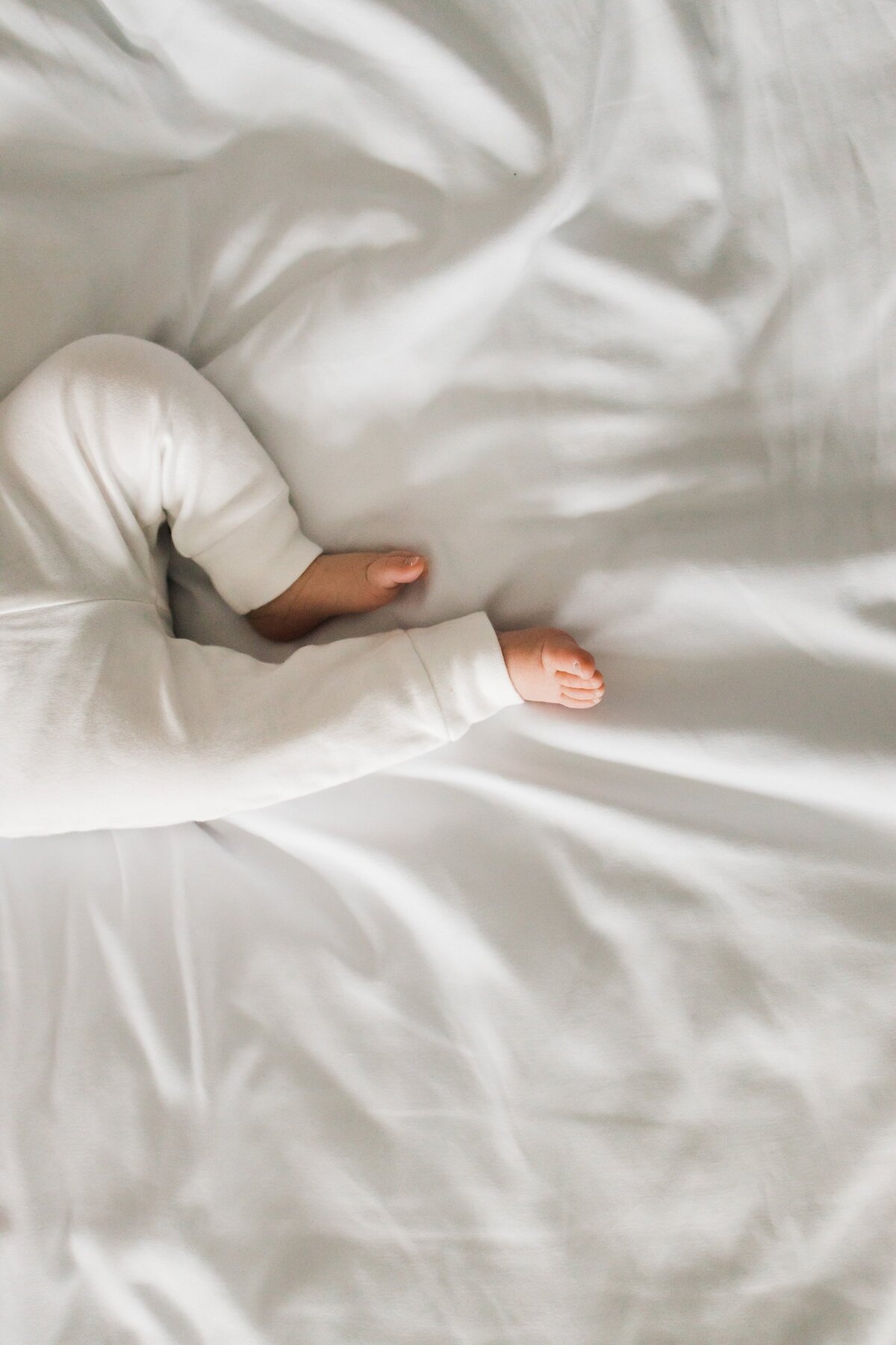 Child in white pajamas lying under white bedcovers with only feet and one arm visible, captured during an in-home newborn photography session.