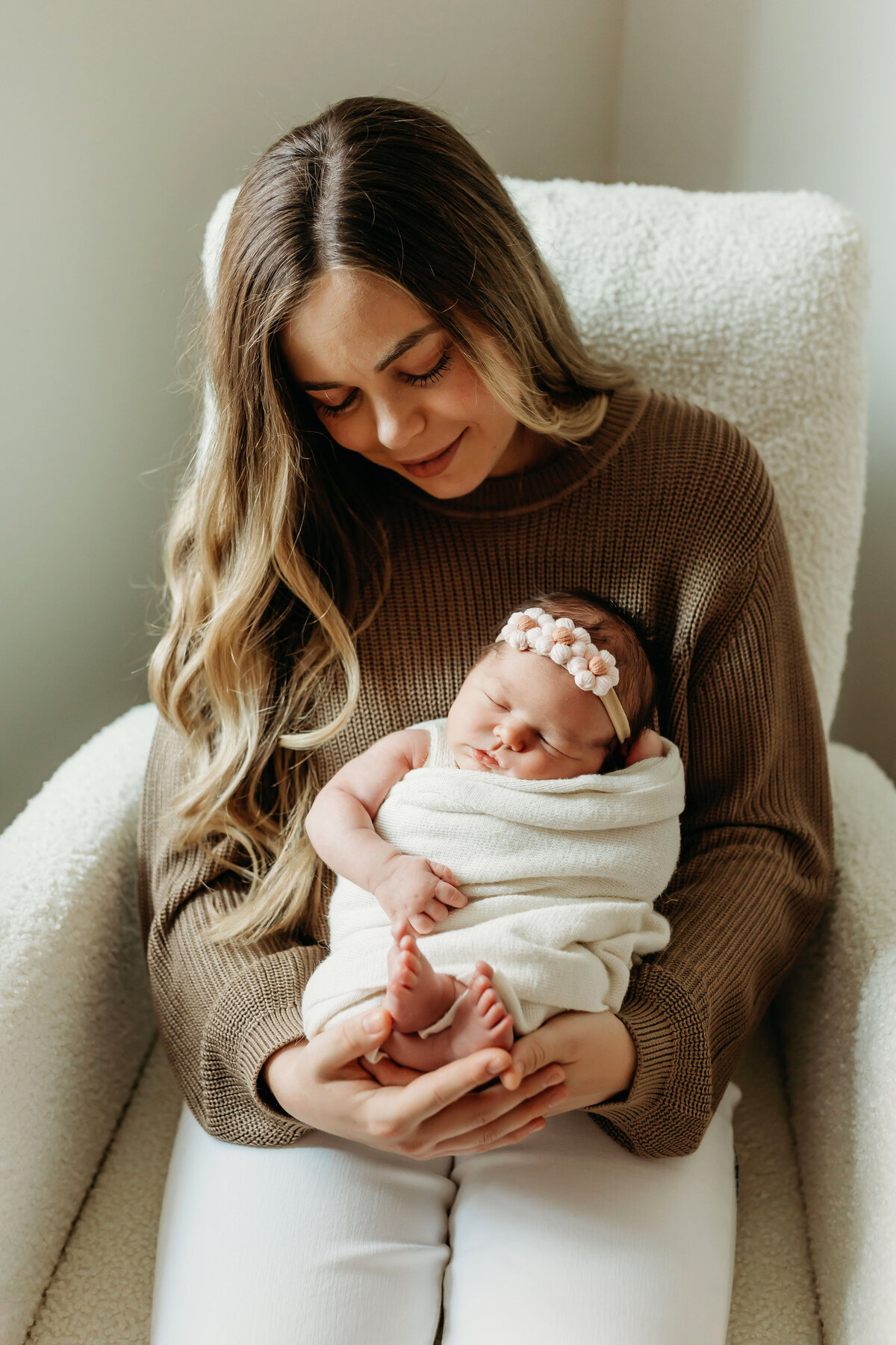 A mom sits in a rocking chair new her nursery window, holding her newborn baby