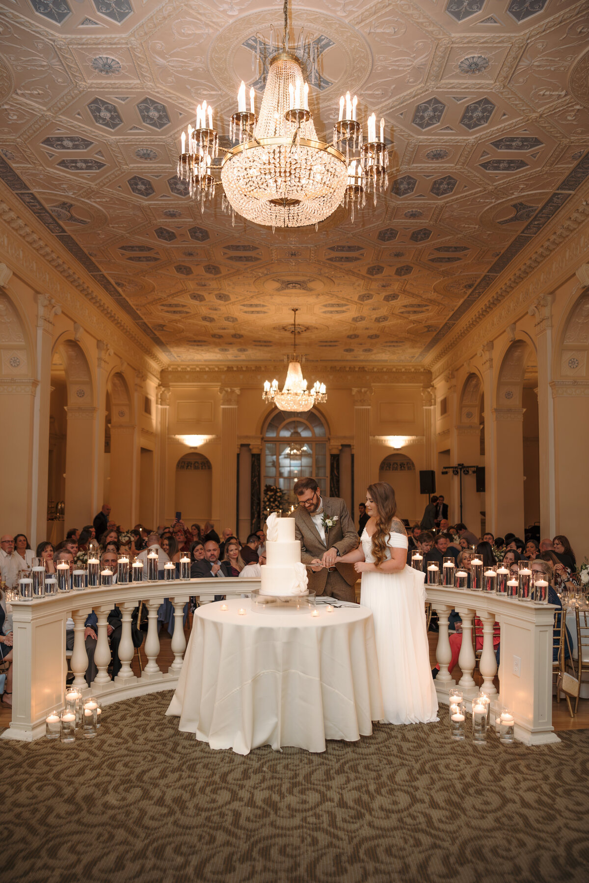 Bride and groom cutting their white tiered wedding cake in a candlelit ballroom.